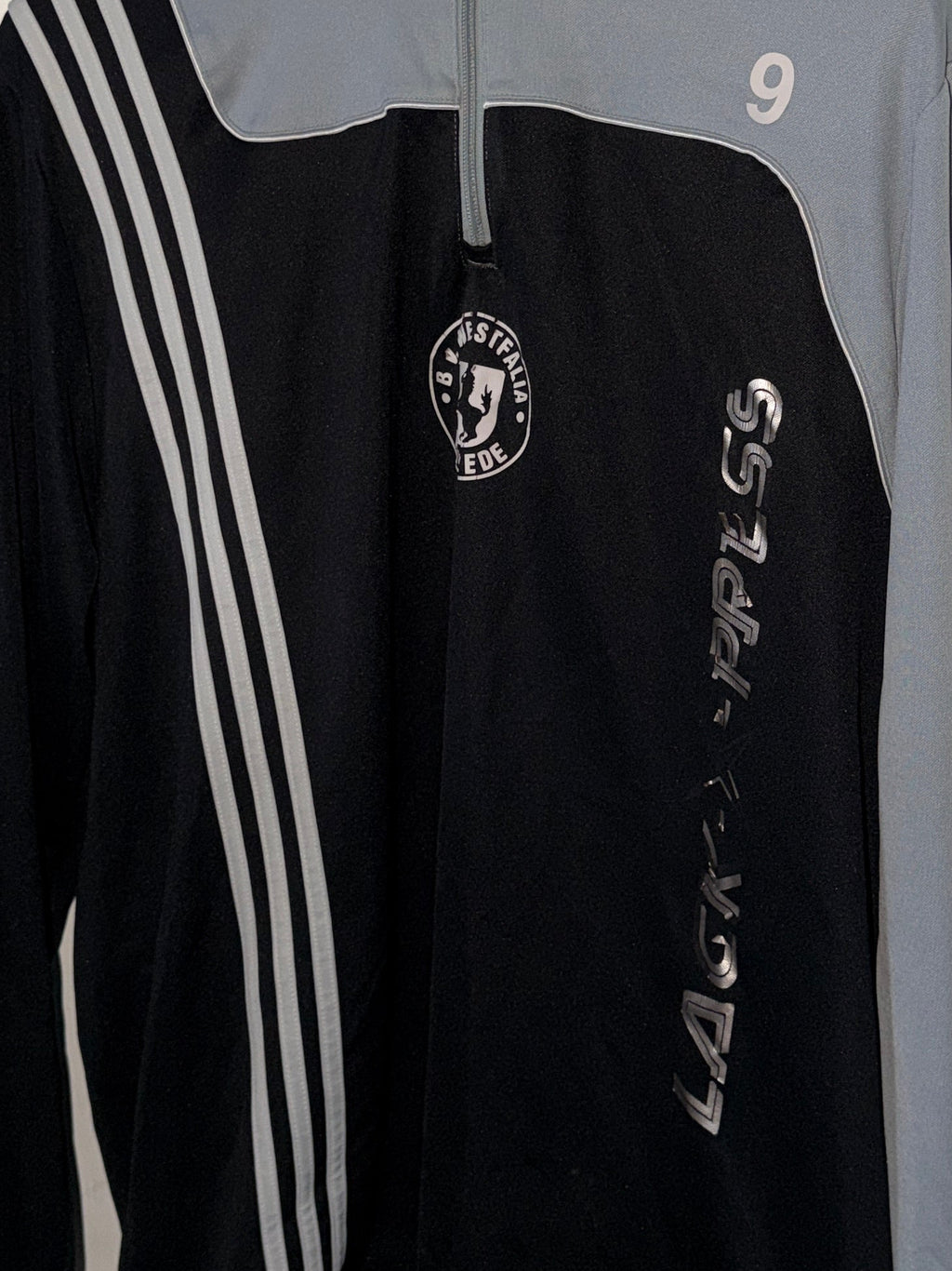 Black sports jacket with white stripes and logo on a hanger against a white wall.