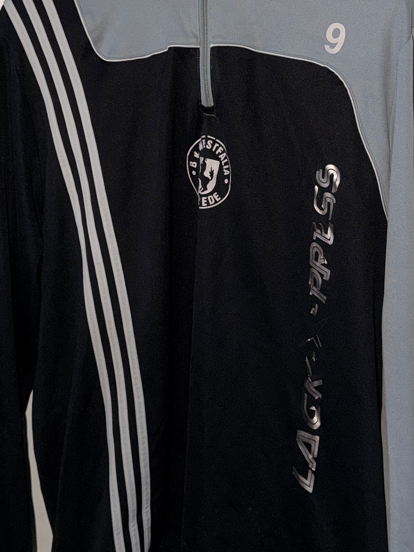 Black sports jacket with white stripes and logo on a hanger against a white wall.