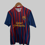 Barcelona football jersey with Qatar Foundation logo on a hanger against a white wall.