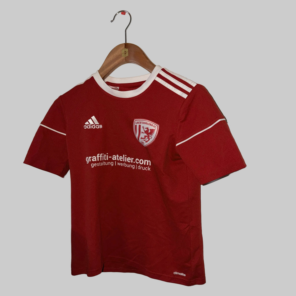 Red sports jersey with Adidas logo on a hanger against a white wall.