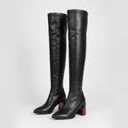 A pair of black leather over-the-knee boots with pointed toes and chunky block heels, featuring a distinctive red sole on the heel, photographed against a light gray studio background.