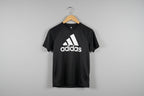Black Adidas t-shirt on a wooden hanger against a textured white wall.